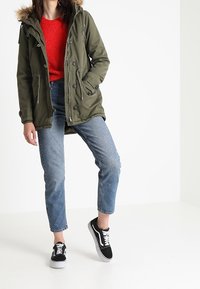 Olivgrön parkajacka med foder av päls, röd tröja och blå korta jeans. Svarta sneakers med vita sulor. Enkel, avslappnad stil.