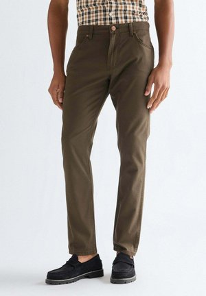 Mann trägt braune gerade geschnittene Hosen, schwarze Loafers und ein in die Hose gestecktes kariertes Hemd, steht vor einem einfarbigen hellen Hintergrund.