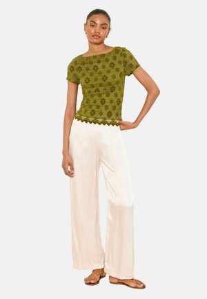 Naine kannab oliivirohelist lühikeste varrukatega heegeldatud topi, valgeid laia säärega pükse ja pruune lameda tallaga sandaale, seistes ühe käega puusal.