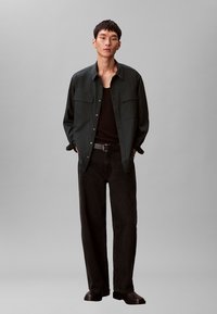 Camicia verde scuro con due tasche frontali, canotta nera e pantaloni larghi e scuri. La modella indossa stivaletti neri lucidi. Design semplice e moderno.