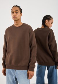 To modeller med fletter iført brune hettegensere og lyseblå jeans, den ene vender mot kameraet og den andre er vendt bort.