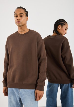 To modeller med fletter iført brune hettegensere og lyseblå jeans, den ene vender mot kameraet og den andre er vendt bort.