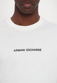 Fehér pamut póló, környakú fazonnal és fekete "ARMANI EXCHANGE" logóval a mellrészén. Sima textúra, laza illeszkedés.