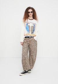 Άσπρο πουκάμισο με μακριά μανίκια, διακοσμημένο με γαλάζιο και πορτοκαλί γραφικό πουλιού, σε συνδυασμό με μπεζ παντελόνι φαρδιάς γραμμής με λεοπάρ σχέδιο και μαύρα αθλητικά παπούτσια.