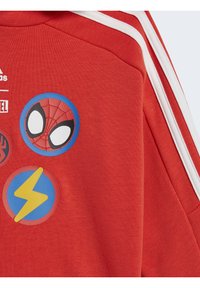 Felpa rossa con strisce bianche. Presenta tre toppe circolari: Spider-Man, un cerchio blu con un fulmine e un emblema di ragno nero.