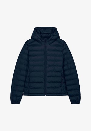 Femme debout les mains dans les poches, portant une veste matelassée à capuche bleu marine, un jean gris foncé ample et des baskets blanches sur un fond uni.