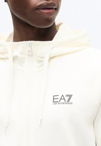 Κοντινό πλάνο ενός ατόμου που φοράει λευκό φούτερ με φερμουάρ και κουκούλα, με το λογότυπο "EA7 Emporio Armani" στο αριστερό μέρος του στήθους και κορδόνια στην κουκούλα.