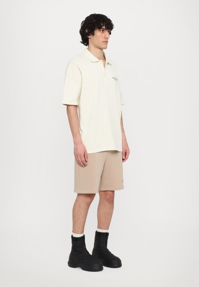 Polo shirt crème avec un col et texte vert, associé à un short beige. Porté avec des bottes zippées noires avec des accents blancs.