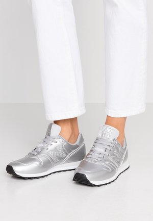 Person iført skinnende sølvfarvede New Balance-sneakers med hvide såler og hvide snørebånd, kombineret med afkortede hvide bukser, står indendørs.
