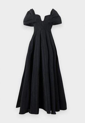 Žena s krátkymi vlasmi v dlhých čiernych šatách bez ramien s veľkými nafúknutými rukávmi a bielymi špicatými podpätkami na bielom pozadí.