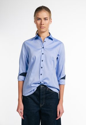 Camicetta azzurro chiaro a maniche lunghe con chiusura a bottoni, caratterizzata da bottoni scuri e polsini arrotolati con un accento blu navy, indossata con pantaloni in denim scuro.