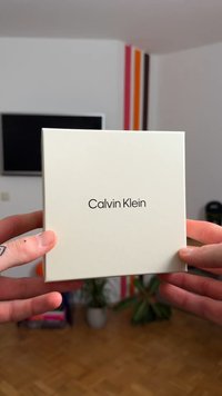 Weißer quadratischer Karton mit dem Schriftzug "Calvin Klein" auf der Vorderseite, in den Händen gehalten. Glatte Oberfläche; minimalistisches Design ohne zusätzliche Grafiken oder Texturen.