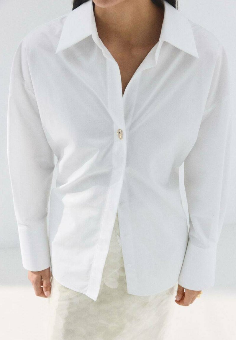 Chemise blanche à manches longues avec un grand col et un seul bouton doré à l'avant. Texture lisse, coupe ajustée et coupe droite.