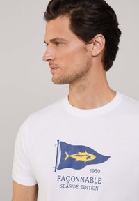 Camiseta blanca de algodón con un gráfico de una bandera azul y un pez amarillo, con el texto "EDICIÓN SEASIDE 1950 FAÇONNABLE" debajo.