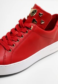 Baskets en cuir rouge avec bout rond, semelle blanche plate, lacets rouges plats et accents dorés en forme de cœur sur la languette.