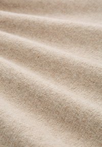 Beżowy materiał tekstylny o miękkiej, fakturowanej powierzchni z delikatnymi odcieniami koloru i tkanym wzorem, układający się w łagodne fałdy.