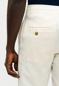 Κοντινή λήψη ενός ατόμου που φοράει παντελόνι λινό σε εκρού χρώμα με κουμπωτή πίσω τσέπη, το χέρι του χαλαρό στο πλευρό.