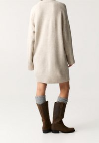 Une personne portant une robe en tricot beige, des chaussettes grises amples et des bottes en cuir marron à mi-mollet, se tenant contre un fond clair et uni.