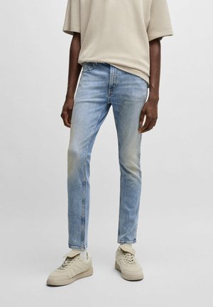 Person trägt hellblaue Slim-Fit-Jeans, beige strukturiertes Kurzarmoberteil und beige Schnürsneaker und steht vor einem schlichten hellen Hintergrund.