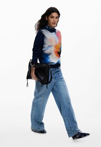 Femeie care merge, purtând un pulover albastru cu flori, blugi largi deschiși la culoare, geantă neagră și adidași cu model, pe un fundal alb simplu.