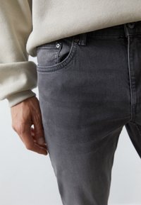 Tmavě šedé džínové kalhoty s přiléhavým střihem, s předními kapsami a jemným vyblednutím. Ruka modelky spočívá blízko otvoru kapsy.