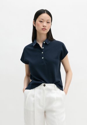 Jonge vrouw staat met handen in de zakken, draagt een marineblauw poloshirt met korte mouwen en witte hooggetailleerde broek tegen een effen achtergrond.