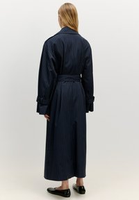 Marinblå trench coat med smalrandig design, breda ärmar, och bälte i midjan, med knälång fåll. Har en klassisk krage och knappdetaljer.