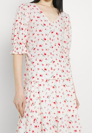 Femme portant un chemisier blanc à col en V et une jupe avec des imprimés floraux rouges et roses, avec des manches bouffantes et un ourlet volanté.