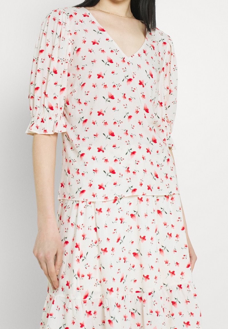 Femme portant un chemisier blanc à col en V et une jupe avec des imprimés floraux rouges et roses, avec des manches bouffantes et un ourlet volanté.