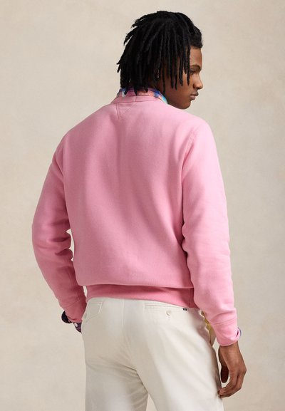 Sweatshirt rosa com acabamento texturizado e punhos canelados. Usada sobre uma camisa xadrez multicolorida, combinada com calças de cor clara.