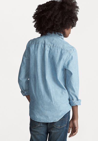 Camisa chambray azul claro com mangas longas, punhos abotoados e bainha curvada. Apresenta detalhes de yoke nas costas e uma textura suave.