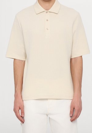 Homme portant un polo beige à manches courtes avec trois boutons et un pantalon blanc, debout devant un fond clair uni.