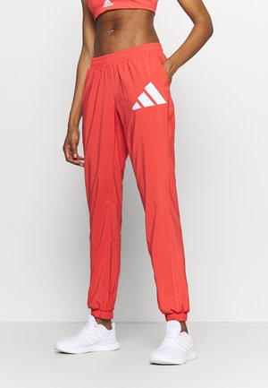 Woman wearing red Adidas track pants and matching crop top with white sneakers, standing with one hand in pocket.