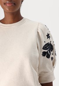 Beige sweatshirt met pofmouwen, voorzien van donkere bloemenborduursels en sterren op de linkerschouder, gemaakt van zacht katoen.