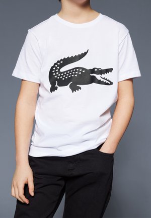 Barn iført en hvid T-shirt med stort sort krokodillemotiv og sorte bukser, den ene hånd i lommen, mod en ensfarvet grå baggrund.