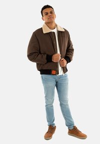 Braune Bomberjacke mit einem cremefarbenen Pelzkragen, schwarzen gerippten Bündchen und einem kleinen orangefarbenen Etikett. Hellblaue Jeans und braune Schuhe runden das Outfit ab.