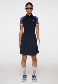 Granatowa koszulka golfowa z jasnoniebieskimi akcentami, krótki rękaw i logo. W połączeniu z granatową spódnicą, szarymi skarpetkami w wzory i niebieskimi butami sportowymi.