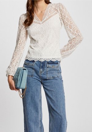 Mujer que lleva una blusa blanca de encaje con mangas largas y dobladillo festoneado, y vaqueros azules de talle alto, sosteniendo un pequeño bolso azul con cadena dorada.