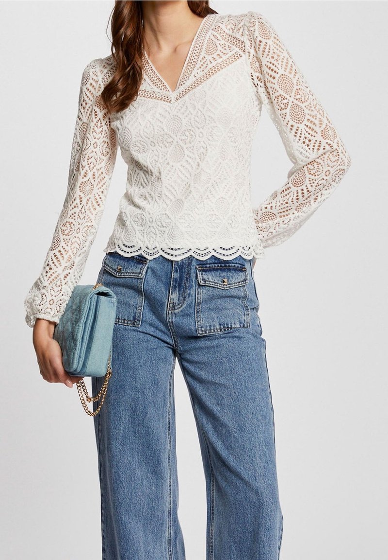 Femme portant un chemisier en dentelle blanche à manches longues avec un ourlet festonné et un jean bleu taille haute, tenant un petit sac bleu avec une chaîne dorée.