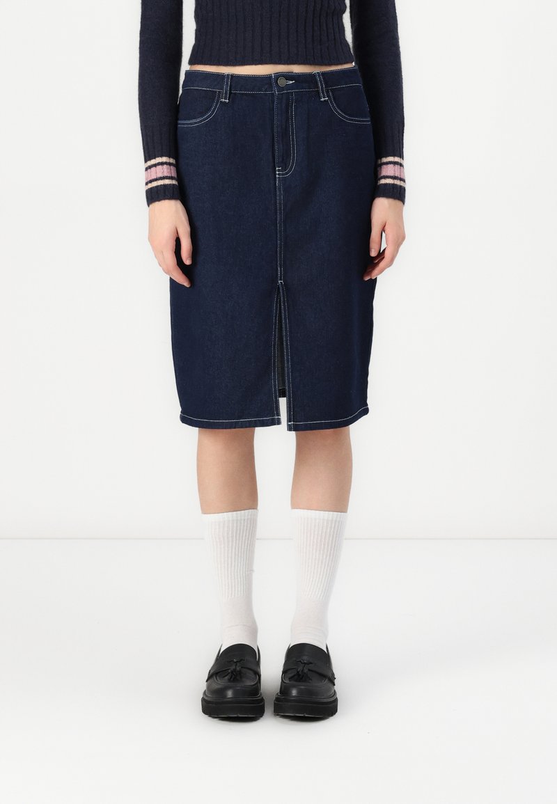 Mørkeblå denim midi skjørt med forgående splitt, hvite sømmer og klassisk fem-lommers design. Kombinert med hvite ribbestrikkede sokker og svarte sko.