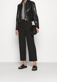 Schwarze, lederne cropped Jacke mit spitzem Kragen, kombiniert mit weiten schwarzen Hosen und schwarzen Rutsch-Sandalen. Clutch-Tasche mit strukturiertem Muster.