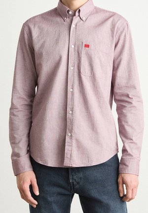 Mann trägt ein helllila Hemd mit Brusttasche und kleinem rotem Logo-Patch, kombiniert mit dunkelblauen Jeans.