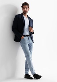 Marineblazer über einem weißen V-Ausschnitt-Shirt, hellblauen Jeans und schwarzen Schuhen. Das Model posiert vor einem weißen Hintergrund, die Hände in den Taschen.