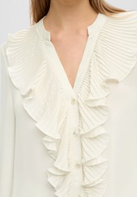 Blusa bianca con rouches plissettate verticali lungo la parte frontale e colletto. Presenta piccoli bottoni e una superficie liscia.