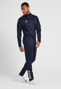 Marine atletiek trainingspak gemaakt van gladde, rekbare stof. Kenmerken zijn een kwart-rits kraag, contrasterende witte schouderstrepen en chevronaccenten op de broek.