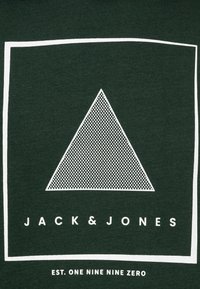 Bílý tečkovaný trojúhelník uvnitř bílého čtvercového rámu na tmavě zeleném pozadí s textem "JACK & JONES" a "EST. DEVĚT NULA" níže.