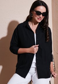Black textured button-up shirt with a collar, featuring a relaxed fit and rolled sleeves, paired with white pants and sunglasses.