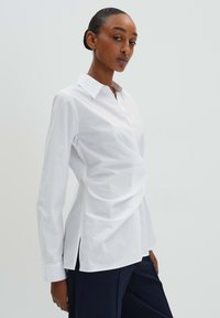 Weißes Langarmhemd mit Kragen, aus einem Stoff mit glatter Textur und mit drapiertem Vorderteil sowie seitlichen Schlitzen. Kombiniert mit dunklen Hosen.
