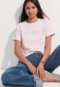 Svijetloružičasta pamučna majica s natpisom „BRUNCH & YAP“ u pisanoj formi, uparena s trapericama visokog struka plave boje i bež teksturiranim obućem.
