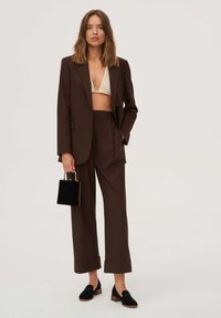 Brązowy garnitur z szerokimi spodniami i oversize'ową marynarką na beżowym dzianinowym topie. Czarne buty na klockowym obcasie i mała czarna torebka dopełniają stylizację.